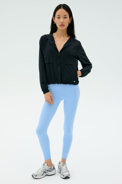 A woman stands against a plain background wearing the SPLITS59 Megan Woven Nylon Jacket in black, paired with light blue spandex leggings and silver athletic shoes.
