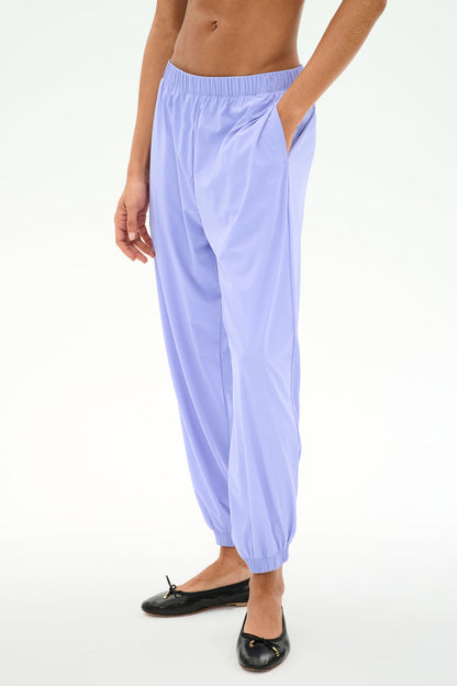 A person wears SPLITS59's Megan Woven Nylon Jogger in Purple Haze, made from stretchy nylon spandex, with hands in pockets and black ballet flats, shown mid-torso down against a plain background.