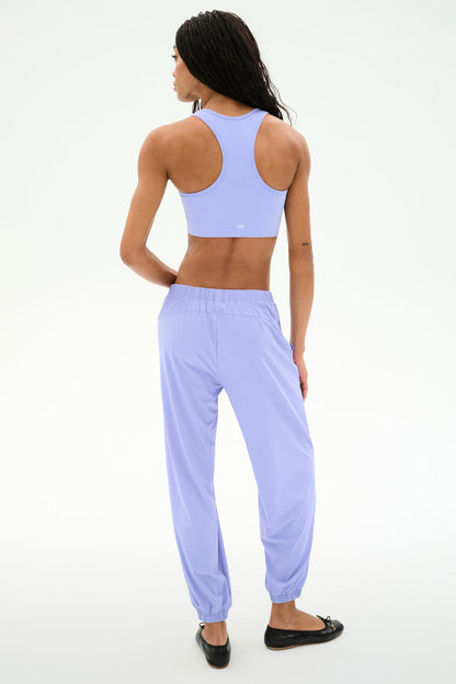 A person with long braided hair faces away from the camera, wearing SPLITS59’s Megan Woven Nylon Jogger in Purple Haze and a matching lavender sports bra, styled with black flats against a plain white background.