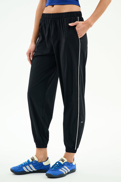 A person stands against a white background wearing SPLITS59 Megan Woven Nylon Jogger w/ Piping - Black/Creme (85% nylon/spandex), featuring a white side stripe, paired with a blue crop top and blue athletic shoes, with one hand in their pocket.