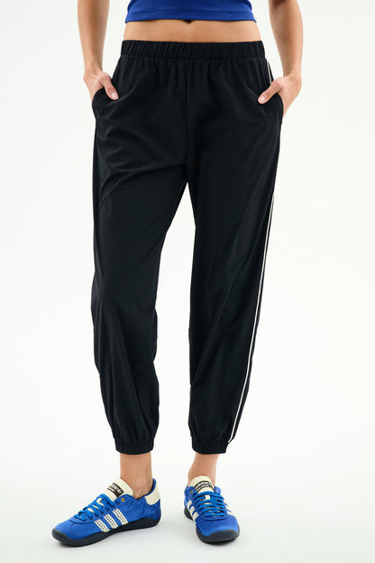 A person wears SPLITS59 Megan Woven Nylon Jogger w/ Piping - Black/Creme, a blue top, and blue athletic shoes, standing with hands in pockets against a plain white background.