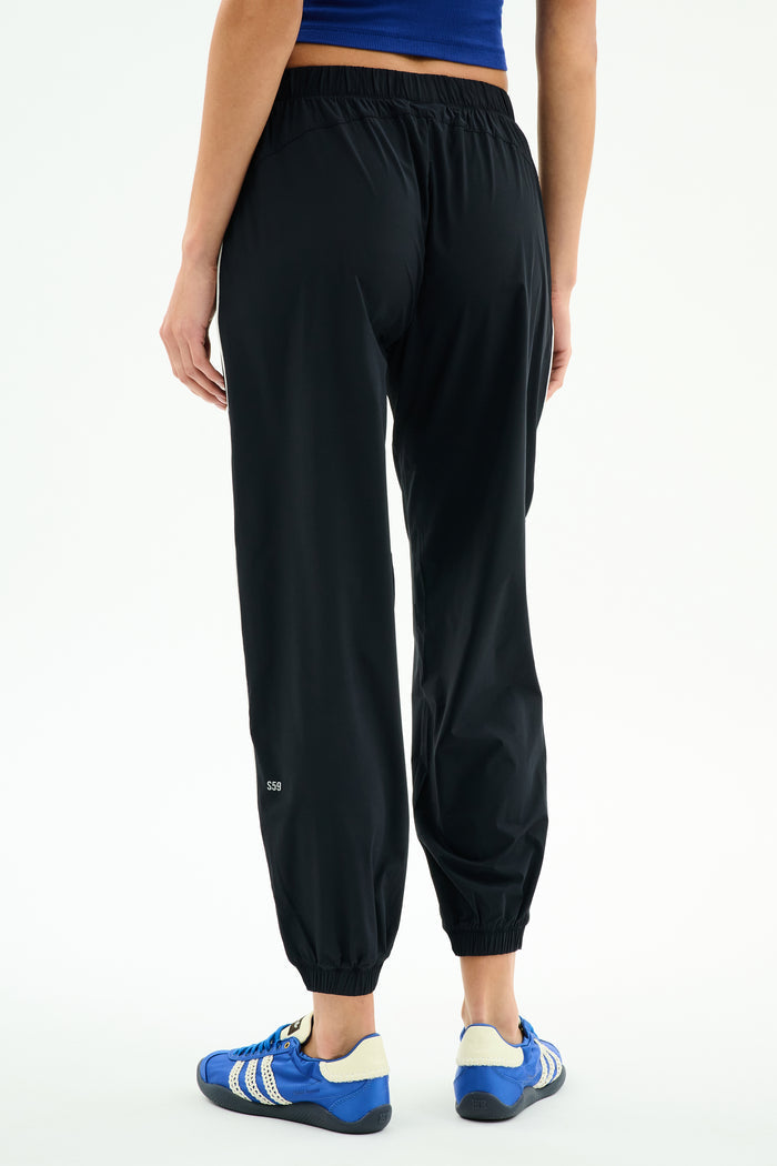 A person wears SPLITS59 Megan Woven Nylon Jogger w/ Piping - Black/Creme, a blue top, and blue athletic shoes, photographed from the back against a plain white background.