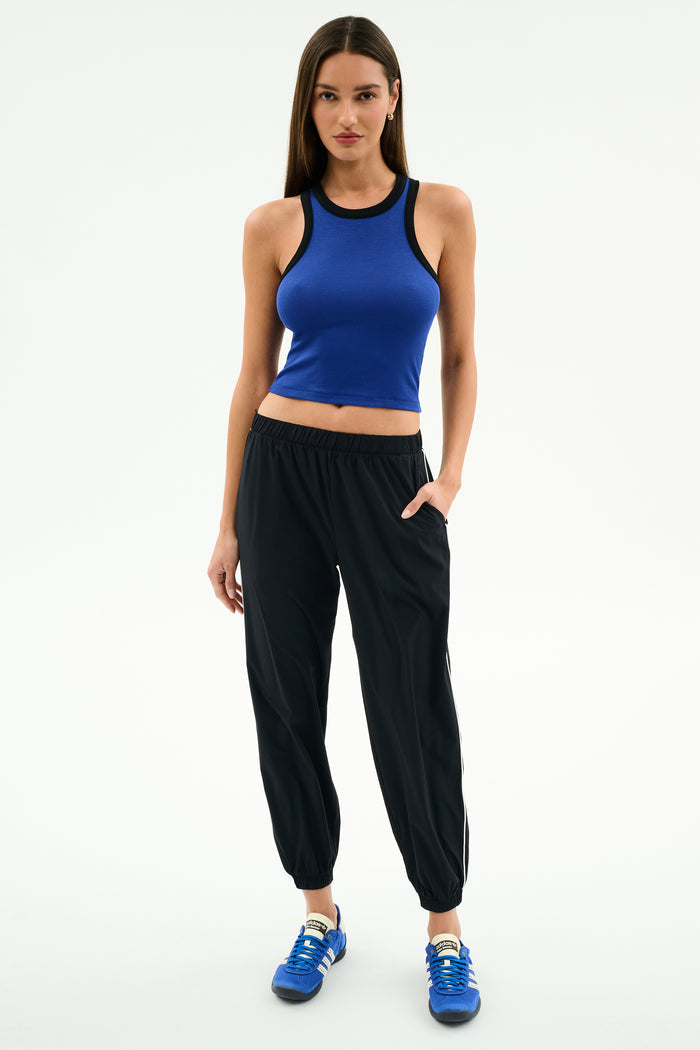 A woman stands against a white background wearing SPLITS59's Megan Woven Nylon Jogger w/ Piping in Black/Creme, paired with a sleeveless blue spandex top and blue sneakers, with one hand in her pocket.