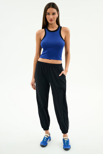 A woman stands against a white background wearing SPLITS59's Megan Woven Nylon Jogger w/ Piping in Black/Creme, paired with a sleeveless blue spandex top and blue sneakers, with one hand in her pocket.