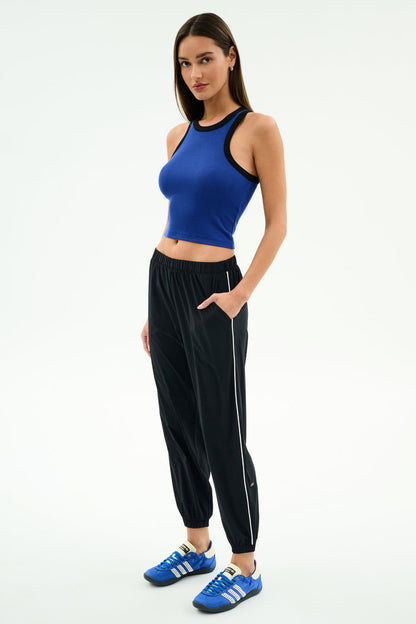 A woman stands against a plain background, wearing a blue sleeveless crop top and SPLITS59 Megan Woven Nylon Jogger w/ Piping in Black/Creme, paired with blue Adidas sneakers.