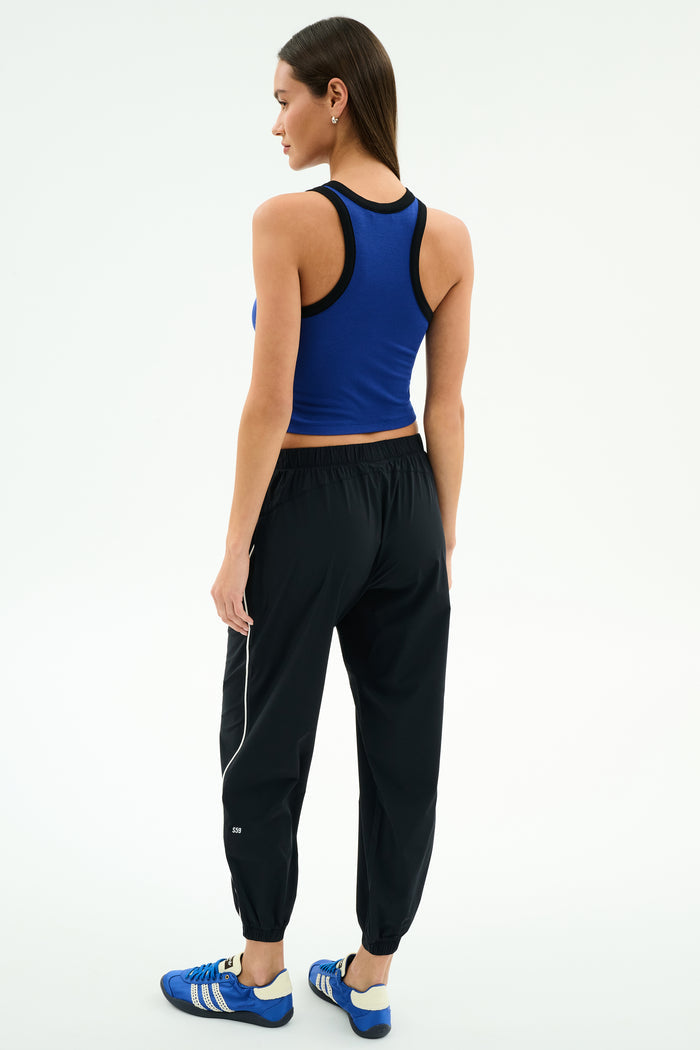 A woman faces away, wearing SPLITS59’s Megan Woven Nylon Jogger w/ Piping in Black/Creme with a blue sleeveless top and blue athletic shoes, crafted from stretchy spandex and 85% nylon, on a plain white background.
