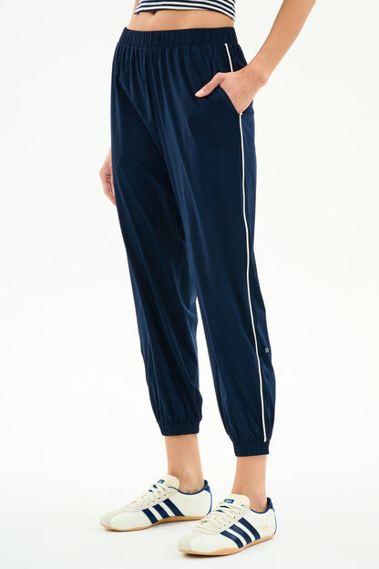 Someone wearing the SPLITS59 Megan Woven Nylon Jogger w/ Piping in Indigo/Creme, styled with a striped top and white Adidas sneakers with navy stripes, standing against a white background.