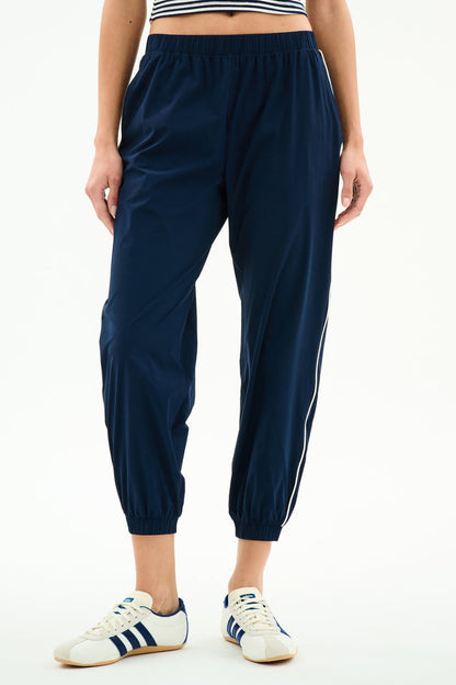 Model wears SPLITS59 Megan Woven Nylon Jogger w/ Piping in Indigo/Creme, featuring a stretchy nylon-spandex blend, paired with white sneakers with blue stripes and a cropped striped top, shown from the waist down against a plain background.