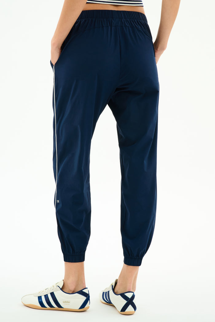 A person models the SPLITS59 Megan Woven Nylon Jogger w/ Piping in Indigo/Creme, featuring white side piping, paired with white Adidas sneakers, standing on a plain white background and photographed from the back.