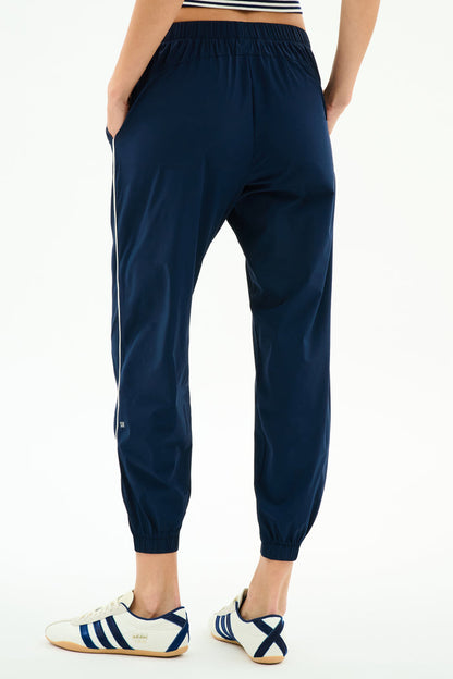 A person models the SPLITS59 Megan Woven Nylon Jogger w/ Piping in Indigo/Creme, featuring white side piping, paired with white Adidas sneakers, standing on a plain white background and photographed from the back.