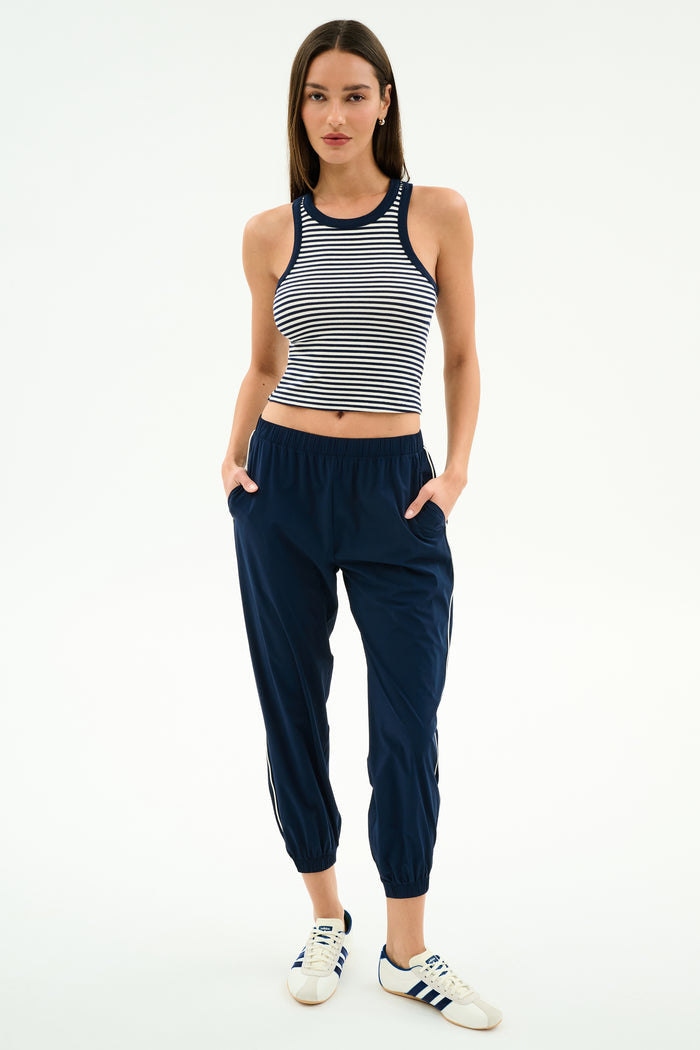 A woman faces forward in a sleeveless striped crop top, SPLITS59 Megan Woven Nylon Jogger w/ Piping in Indigo/Creme, and white Adidas sneakers with blue stripes, posed against a plain white background.