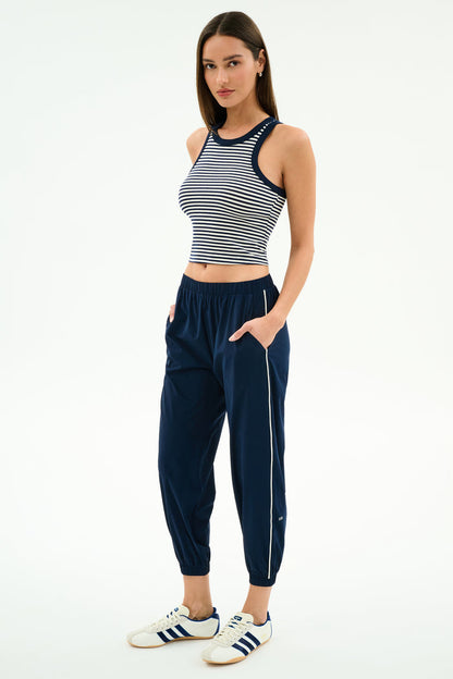 A woman stands against a white background, wearing SPLITS59's Megan Woven Nylon Jogger w/ Piping in Indigo/Creme with a sleeveless striped crop top and white Adidas sneakers. She looks at the camera with her hands in her pockets.