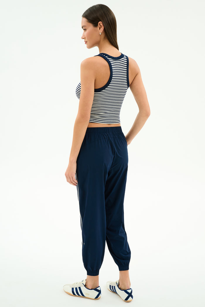 A woman stands slightly turned away, wearing the SPLITS59 Megan Woven Nylon Jogger w/ Piping in Indigo/Creme, a striped sleeveless crop top, and white sneakers with navy stripes against a plain background.