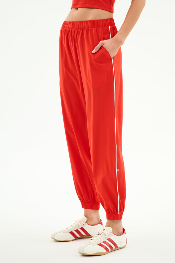 A person wears SPLITS59 Megan Woven Nylon Jogger w/ Piping in Lotus Red/Creme, styled with white sneakers featuring red stripes, and stands with one hand in the pocket against a plain background.