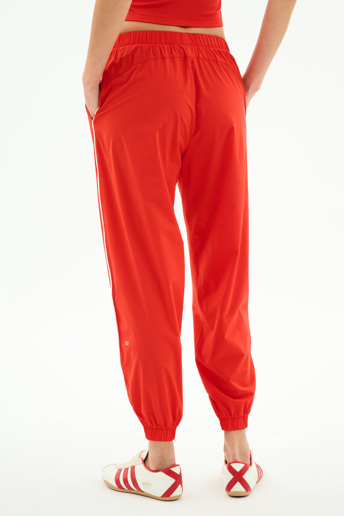 Wearing SPLITS59 Megan Woven Nylon Jogger w/ Piping in Lotus Red/Creme, paired with white sneakers with red accents and a red top, person stands hands in pockets, facing away.