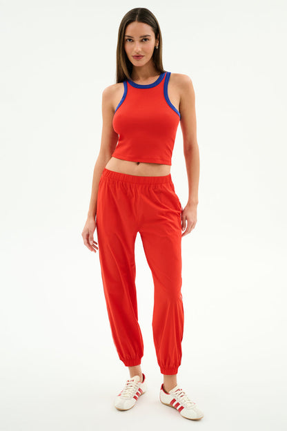 A woman stands against a white background wearing the SPLITS59 Megan Woven Nylon Jogger w/ Piping in Lotus Red/Creme and white sneakers with red stripes.