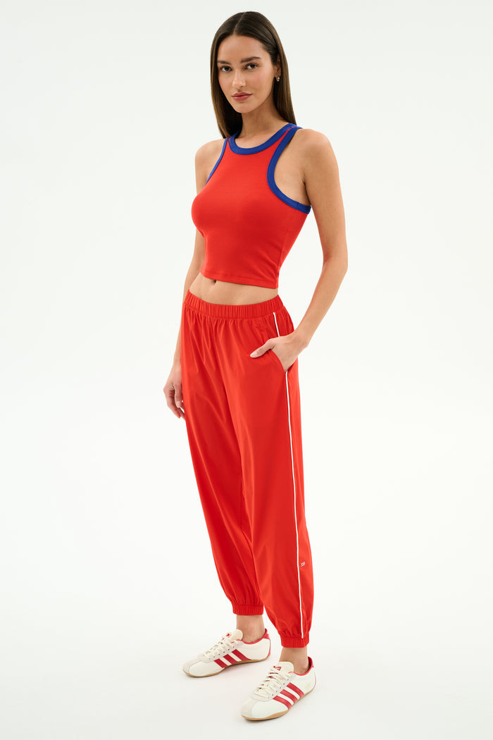 A woman stands against a white background wearing SPLITS59’s Megan Woven Nylon Jogger w/ Piping in Lotus Red/Creme, paired with a red sleeveless crop top and white sneakers with red stripes.