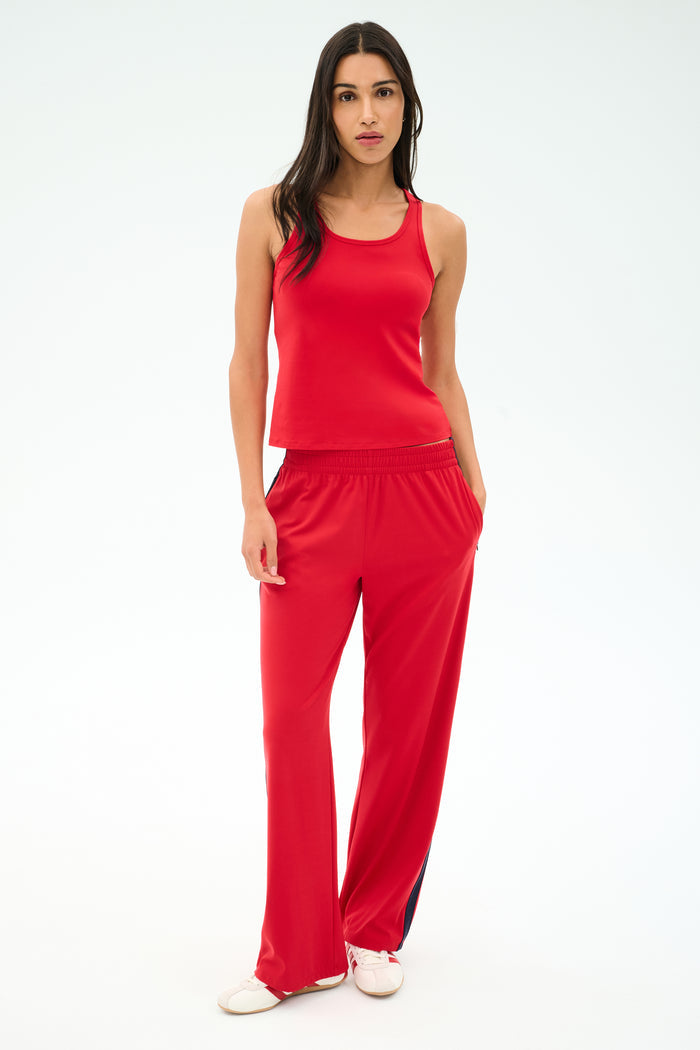 A woman faces forward in a SPLITS59 Mel Airweight Lite Tank in Pirate Red, paired with matching red pants with side stripes and white sneakers, set against a plain light backdrop.