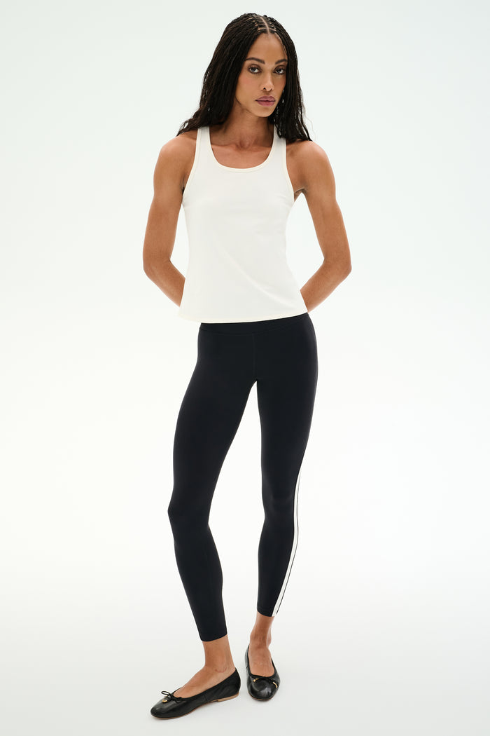 A woman poses against a white background in the SPLITS59 Mel Airweight Lite Tank - White, paired with black side-stripe leggings and black ballet flats—ideal for Pilates or hot yoga.
