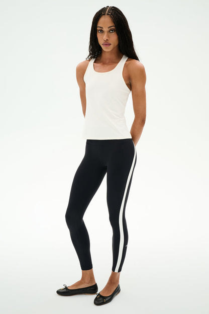 Person wearing the SPLITS59 Mel Airweight Lite Tank in white, perfect for hot yoga or Pilates, paired with black leggings with a white side stripe and black flats against a white background.