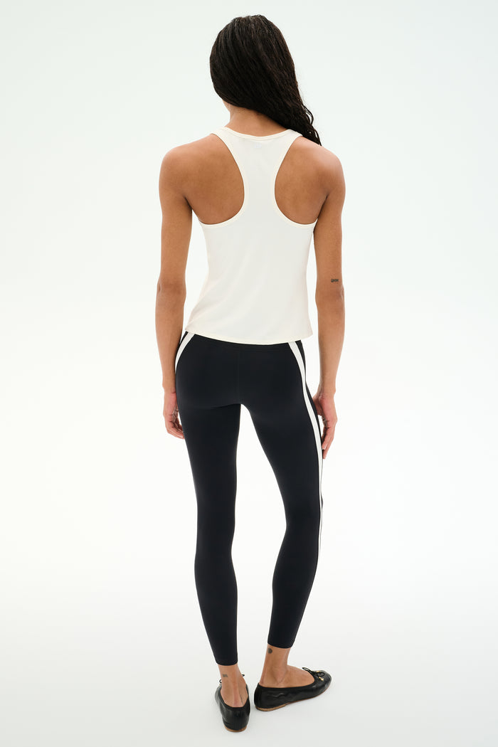 A person stands on a white background, facing away, wearing the SPLITS59 Mel Airweight Lite Tank in white—ideal for Pilates—paired with black leggings featuring white side stripes and black flats.