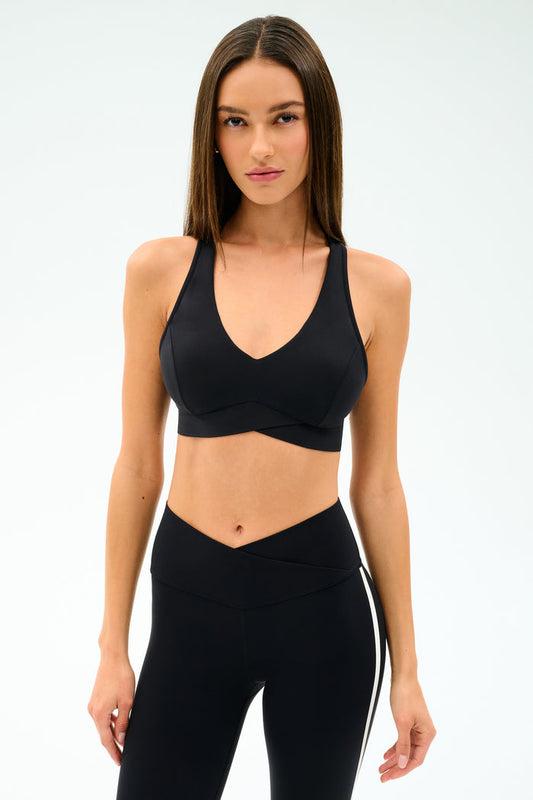 A woman wears the SPLITS59 Mia Rigor Bra in black with high-waisted black leggings, standing against a plain white background—perfect for gym workouts and multi-sport performance.
