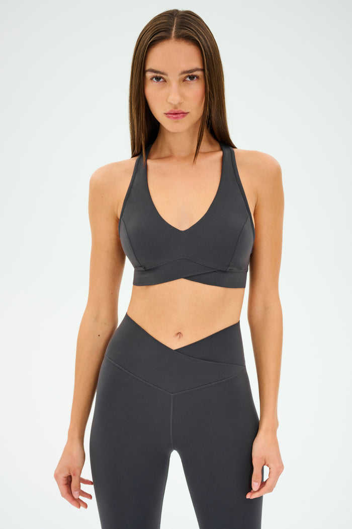 A woman stands facing forward against a plain white background, wearing the SPLITS59 Mia Rigor Bra in Graphite paired with matching high-waisted leggings, both designed for multisport performance.