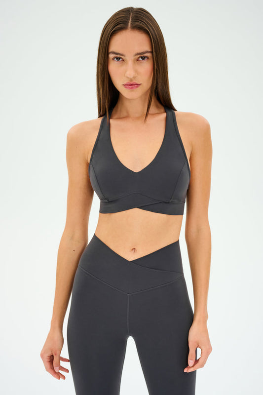 A woman stands facing forward against a plain white background, wearing the SPLITS59 Mia Rigor Bra in Graphite paired with matching high-waisted leggings, both designed for multisport performance.