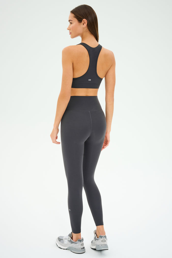 A woman stands on a white background, facing away, wearing the SPLITS59 Mia Rigor Bra in Graphite—a black racerback bra designed for multi-sport performance—paired with matching high-waisted leggings and gray sneakers, making it perfect for gym workouts.