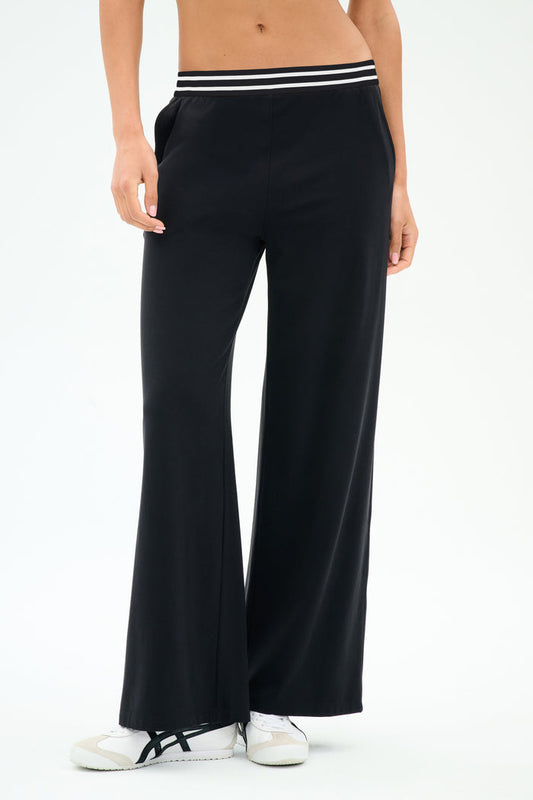 A person wears SPLITS59 Michele Airweight Trouser - Black, featuring a wide-leg fit and a white-and-black striped waistband, paired with white sneakers against a plain background.