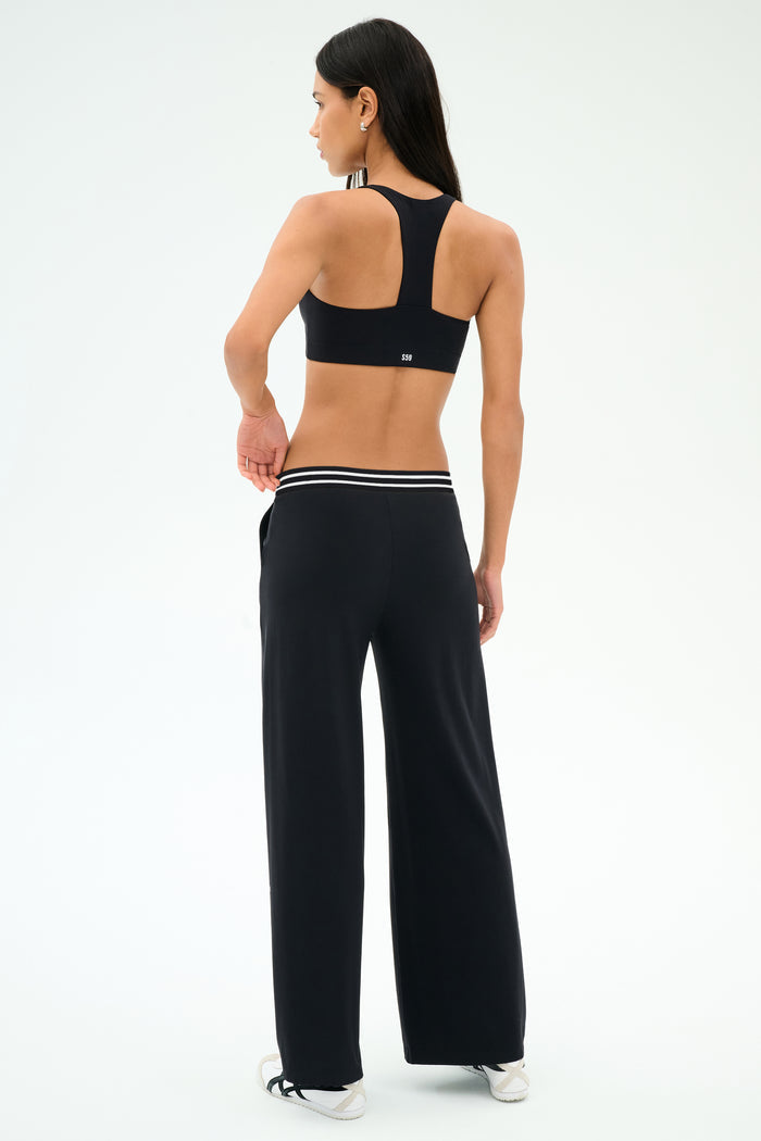 A woman stands facing away, wearing a black sports bra, white sneakers, and SPLITS59 Michele Airweight Trouser - Black with a striped waistband, posed against a white background.