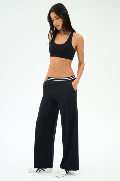 A woman stands with hands in pockets against a plain white background, wearing a black sports bra, SPLITS59 Michele Airweight Trouser - Black with a striped waistband, and white sneakers.