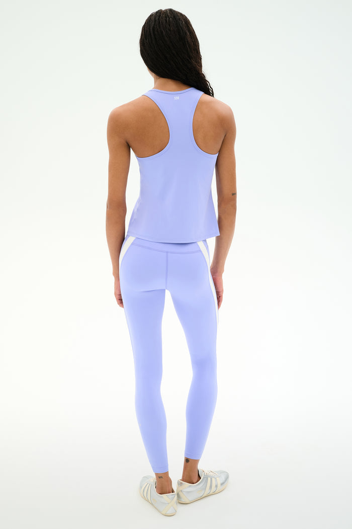 A person stands facing away, wearing SPLITS59 Miles High Waist Rigor 7/8 leggings in Purple Haze/White, styled with a light purple athletic tank top and white sneakers against a plain white background.
