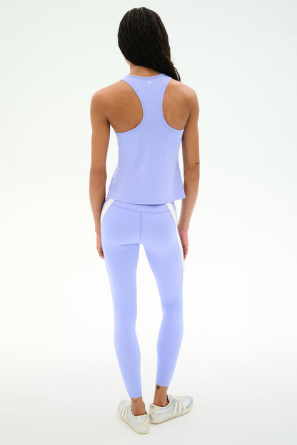 A person stands facing away, wearing SPLITS59 Miles High Waist Rigor 7/8 leggings in Purple Haze/White, styled with a light purple athletic tank top and white sneakers against a plain white background.