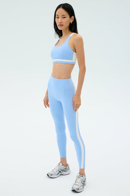 A woman stands on a plain background wearing the SPLITS59 Miles High Waist Rigor 7/8 leggings in Skylight/White and a matching sports bra, paired with silver athletic shoes.