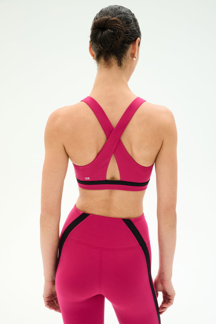 Woman with dark hair in a bun wears the SPLITS59 Miles Rigor Bra in Sangria/Black, featuring crisscross straps and matching leggings. Both are made from a soft nylon spandex blend, shown from the back against a plain background.