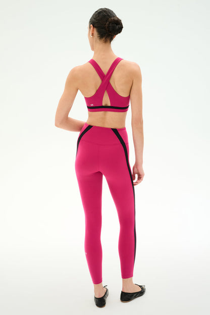 A woman faces away, wearing the SPLITS59 Miles Rigor Bra in Sangria/Black with crisscross straps and matching leggings with black side stripes, both made from sleek nylon spandex, set against a plain white background.