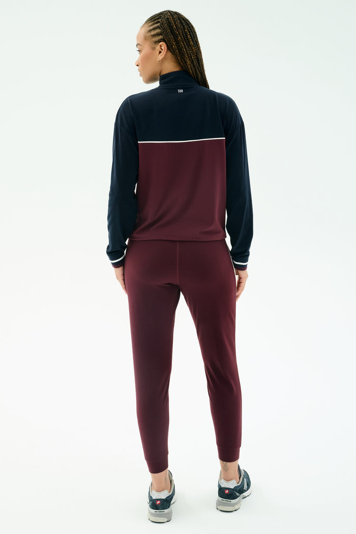 A person with braided hair faces away, wearing the SPLITS59 Millie Airweight Half Zip in Dark Cherry/Indigo and athletic shoes, shown against a plain white background.