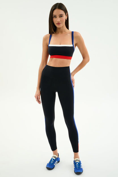 A woman stands against a white background wearing a red, white, and blue striped sports bra, SPLITS59 Monah High Waist Rigor Legging 7/8 in Black/Cobalt, and blue sneakers.