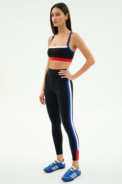 A woman wears SPLITS59's Monah High Waist Rigor Legging 7/8 in Black/Cobalt with blue, white, and red stripes and a matching black sports bra, paired with blue Adidas sneakers against a plain white background.