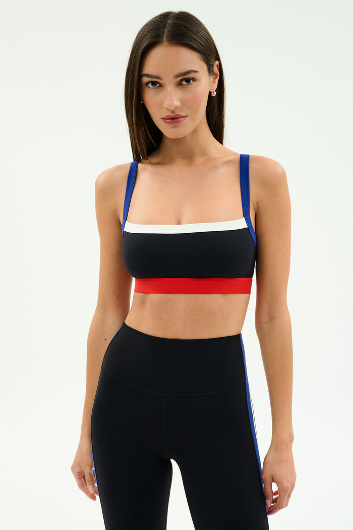 A woman models the SPLITS59 Monah Rigor Bra in Black/Cobalt with matching high-waisted leggings, standing against a plain, light background.