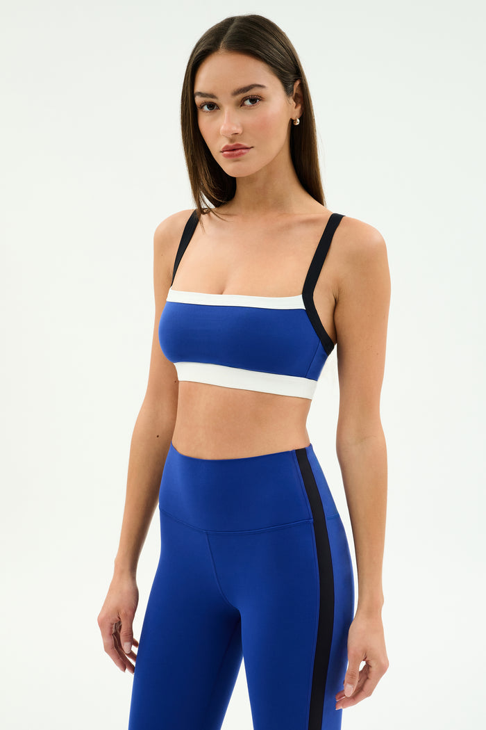 A woman stands against a plain background wearing the SPLITS59 Monah Rigor Bra in Cobalt/Black, paired with matching high-waisted blue leggings featuring black side stripes.