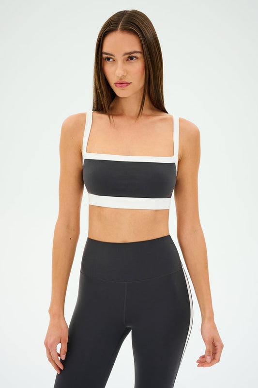 Poised against a plain backdrop, a person wearing a chic SPLITS59 Monah Rigor Bra in Graphite/White and black leggings is ready to conquer their gym workouts.
