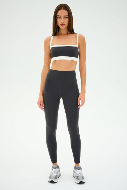 A person stands in a gray and white athletic outfit, ready for gym workouts. The sleek design of the SPLITS59 Monah Rigor Bra - Graphite/White adds style against the plain background.