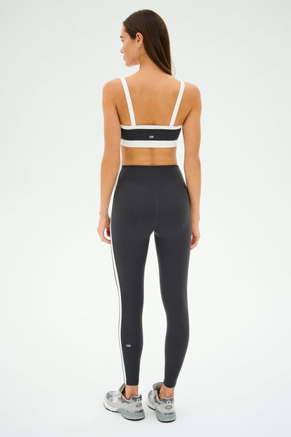 Facing away on a light background, someone wears a SPLITS59 Monah Rigor Bra in Graphite/White, paired with black leggings and gray sneakers, ready for gym workouts.
