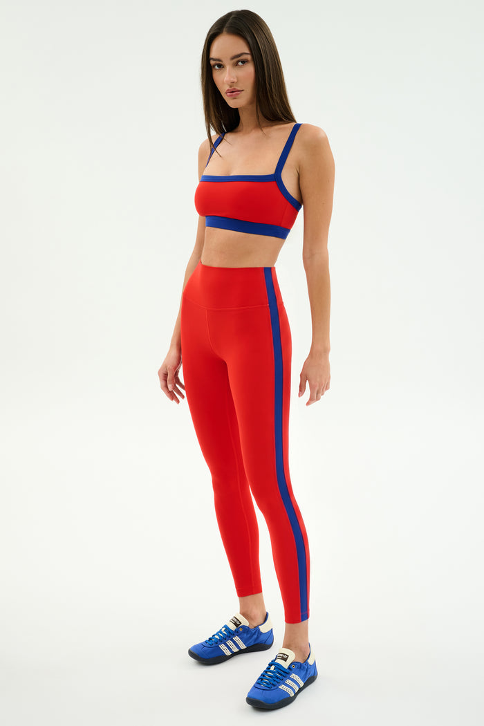 A woman models the SPLITS59 Monah Rigor Bra in Lotus Red/Cobalt, paired with matching high-waisted leggings and blue athletic shoes, standing confidently against a plain white background.