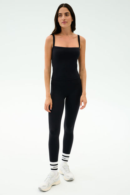 A woman stands against a plain background wearing the SPLITS59 Monah Rigor Tank in black, paired with black leggings, white athletic shoes, and white socks with black stripes—ideal for gym workouts.