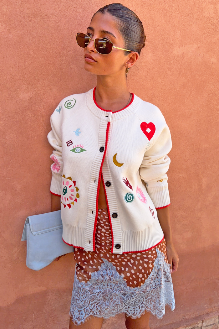 Morocco Charms Cheryl Cardigan in cream with red trim and embroidered motifs, styled on a model with a blue clutch bag.