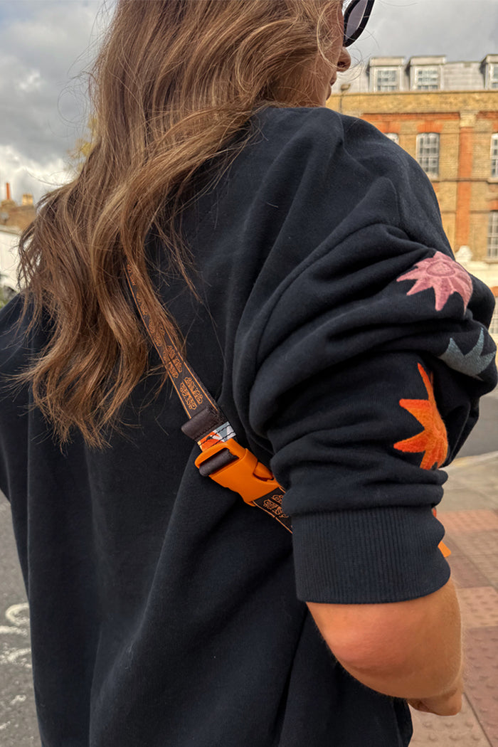 Navy Charm Bumbag with adjustable charm print strap and orange buckle, styled over a navy motif sweatshirt in London.