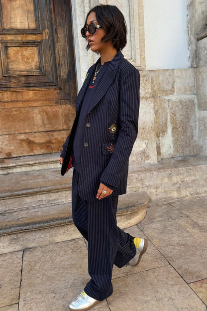 Navy Pinstripe Arden Trousers and matching blazer with sun and eye embroidery, styled with silver trainers.